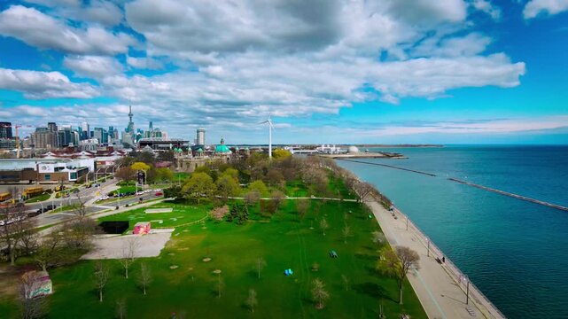 Cinematic aerial drone footage of Marilyn Bell Park along Lake Ontario in Toronto, Canada. The scene captures a vibrant lakeside walking path with pedestrians, light urban traffic, and sweeping views 