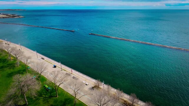 Cinematic aerial drone footage of Marilyn Bell Park along Lake Ontario in Toronto, Canada. The scene captures a vibrant lakeside walking path with pedestrians, light urban traffic, and sweeping views 