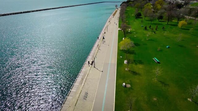 Cinematic aerial drone footage of Marilyn Bell Park along Lake Ontario in Toronto, Canada. The scene captures a vibrant lakeside walking path with pedestrians, light urban traffic, and sweeping views 