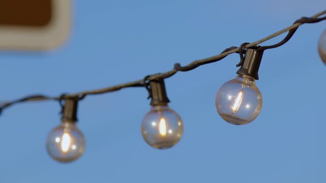 Row of spherical light bulbs, suspended on a wire, emits a warm, soft glow against the backdrop of a clear blue sky. The scene conveys a cozy, festive atmosphere, perfect for outdoor evening events or