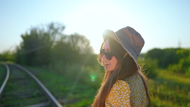 Happy woman in hat stands near railroad tracks at sunset. Smiling girl in sunglasses looks back enjoying summer sun. Female traveler by railway track. Person waits for train in sunny nature.