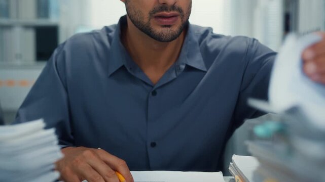 Closeup overworked young Latin businessman reading document in large stack of paperwork file at office desk. Tax audit deadline, accounting stress, corporate burnout, bookkeeping, bureaucracy concept.
