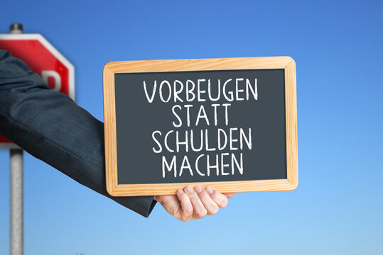 Schultafel mit der Botschaft &bdquo;VORBEUGEN STATT SCHULDEN MACHEN&ldquo;, Stoppschild im Hintergrund.