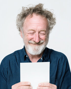 A cheerful elderly man with curly hair and a beard smiles while looking at a blank sheet of paper against a plain background.