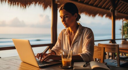 Obraz premium A young woman working on her laptop at a tropical beach cafe during a beautiful golden hour sunset, representing the digital nomad lifestyle and remote work freedom.