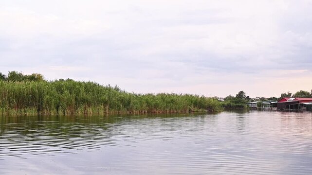 view of boat stations with garages on the lake&rsquo;s shore. Tall reeds surround the area, and the water reflects the cloudy sky, creating a peaceful, rural scene.