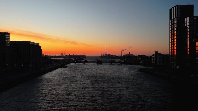 The river flows beneath a vivid sunrise, highlighting Dublin's skyline and distant smokestacks. Ideal for urban landscape projects or travel content.