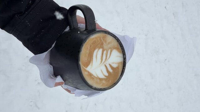 Person holding up a cup of latte in the snow. Light feathery snowflakes blowing over the hot beverage.