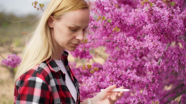 Female agribusiness professional inspecting a blooming orchard tree while using a tablet to manage crop quality, production planning, and agricultural operations.