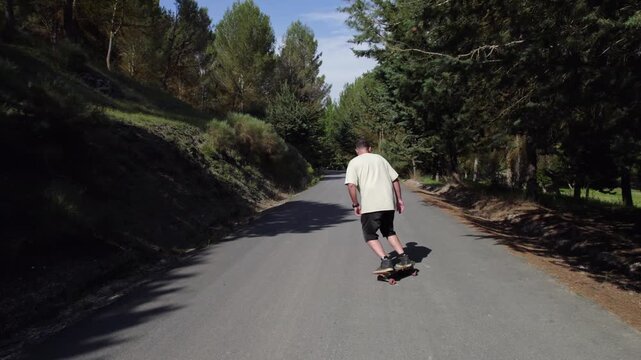 Back view of a mid adult man riding his cruiser skateboard downhill on an empty asphalt road surrounded by a lush green forest on a sunny day	