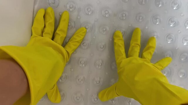 A housekeeper wearing yellow rubber gloves carefully places a transparent non-slip bath mat into a bathtub and secures it using suction cups. everyday home maintenance and hygiene preparation 