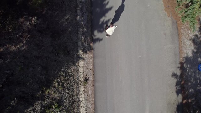 Unrecognizable man riding a cruiser skateboard on an empty road surrounded by trees and vegetation. Aerial top down tracking shot	