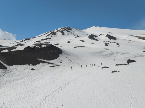 Aerial view of Mount Etna south side with hikers walking on the snow-covered volcanic slopes under a clear blue sky in Nicolosi, Sicily, Italy.