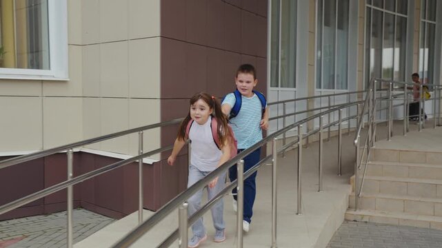 Children are walking along the ramp, School bags are on their shoulders, The building has glass windows, The handrails provide safety, The girl is in front, The boy is following her, The third child