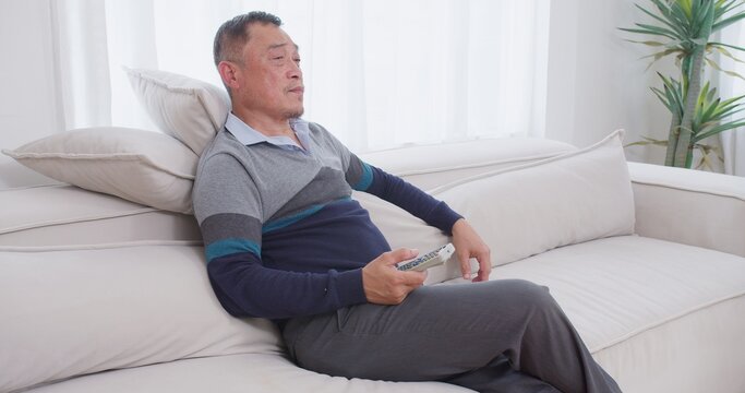 A senior man feeling bored sits on the couch, channel surfing with a remote control while unwinding alone in his bright living room during his retirement.
