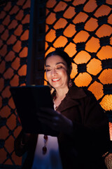 Smiling Hispanic woman reading tablet by decorative window at night, enjoying warm tech-lit atmosphere with focused, pleasant expression of digital interaction. © BullRun