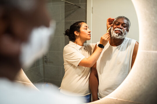 Home caregiver helping senior man shave in bathroom mirror