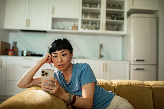 Thoughtful woman on sofa using smartphone in home kitchen