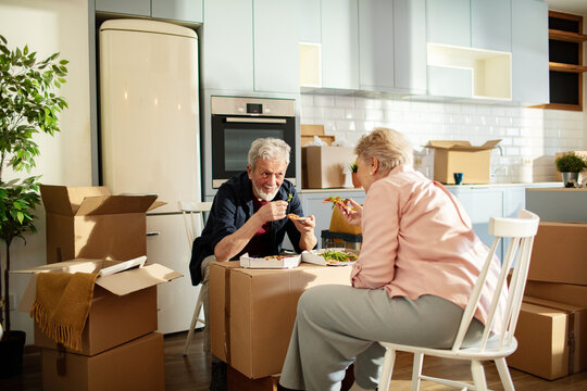 Senior couple eating pizza amid moving boxes in new kitchen