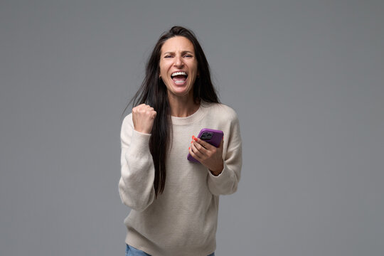 Joyful expressive woman screaming with happiness holding smartphone and clenching fist