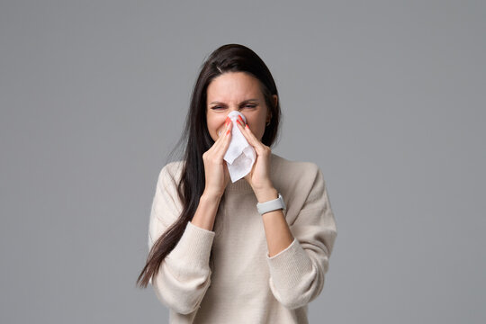 Sick woman in white sweater sneezing into paper tissue isolated on grey background