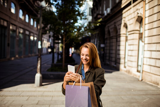 Smiling woman with shopping bags using smartphone on city street