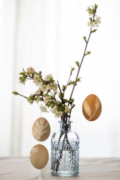 Blown out Easter eggs hang from blossoming branches that stand in a light blue vase against a light background