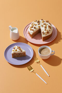 Carrot cake dessert display with coffee, milk and pastel tableware on an orange surface with strong shadows.