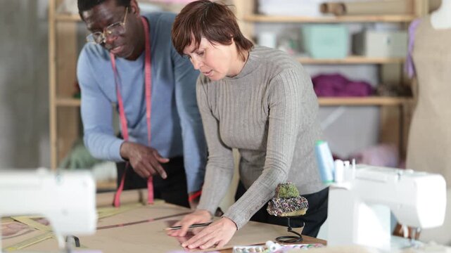 Woman tailor draw dress pattern on paper and participate in creating pattern with African man companion. Craftsman perform paper prototype of product parts, drawing paper pattern for dressmaking.