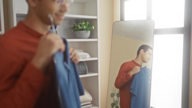 Man holding blue polo shirt on hanger, smiling and presenting the garment in a clothing store fitting room building, shelves visible; contentment.