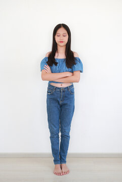 Young Asian girl in blue off-the-shoulder top and denim jeans standing with her arms crossed and eyes closed against white wall background.