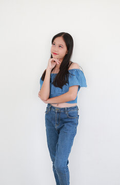 Young girl in blue off-the-shoulder denim top and matching jeans, stands on white background, resting her chin on her hand and looking upward with a curious expression.
