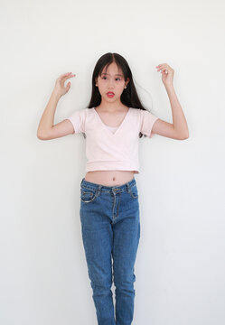 Portrait of young Asian girl in pink top and blue jeans posing playfully while looking beside against white background.