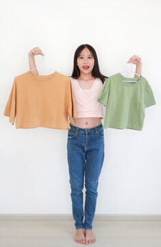 Happy young Asian girl holding two colorful crop top t-shirts on hangers, comparing choices for her daily outfit in bright room.