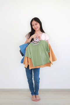 Smiling young Asian girl standing against white wall, happily holding variety of colorful t-shirts on hangers, ready to choose her outfit for the day. Focus at t-shirts in hands.