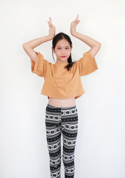 Charming young Asian girl in stylish mustard yellow crop top and patterned leggings posing playfully with her hands over her head against white wall.
