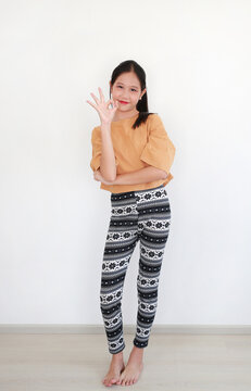 Young Asian girl standing in casual orange t-shirt and patterned leggings, making an "okay" sign with her hand against a white wall.