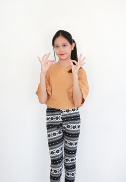 Cheerful Asian young girl in mustard-colored top and patterned leggings posing with a double "OK" hand gesture against white background.