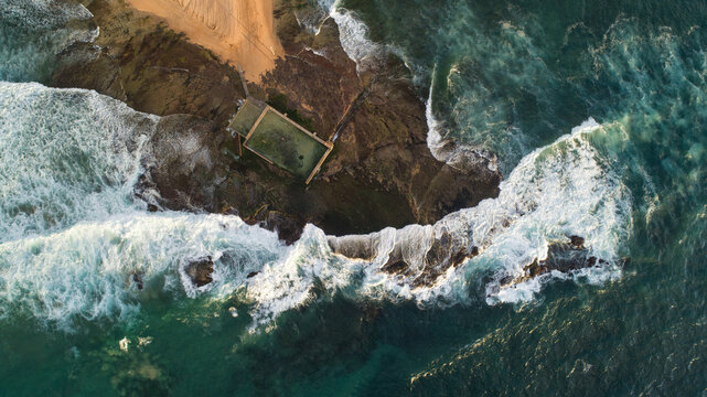 Aerial view of Newport Rock Pool surrounded by crashing waves and white foam on the rocky coastline in Sydney, New South Wales, Australia.