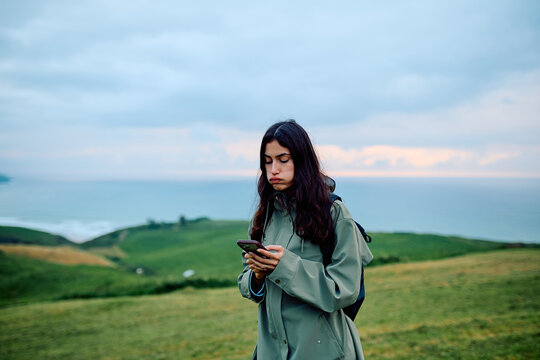 Young woman standing on a green hill overlooking the sea, feeling bored and distracted while using her mobile phone