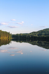 Obraz premium Glassy water reflects forested hills and soft clouds at Ogontz Lake in New Hampshire’s White Mountains near Camp Ogontz at golden hour.