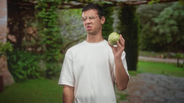 Man holding a custard apple in his right hand, wearing a white t shirt and glasses, grimacing and squinting while inspecting the fruit in a forest; disgust.