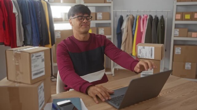 Man wearing glasses and burgundy sweater reaches for laptop with hand among stacked shipping boxes and clothing racks in a small business packing building; small business fatigue.