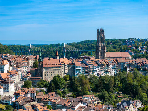 Aerial view of St-Nicolas Cathedral and the historic old town with red-tiled rooftops, the modern Poya Bridge, and lush green hills under a clear blue sky in Fribourg, Fribourg, Switzerland.