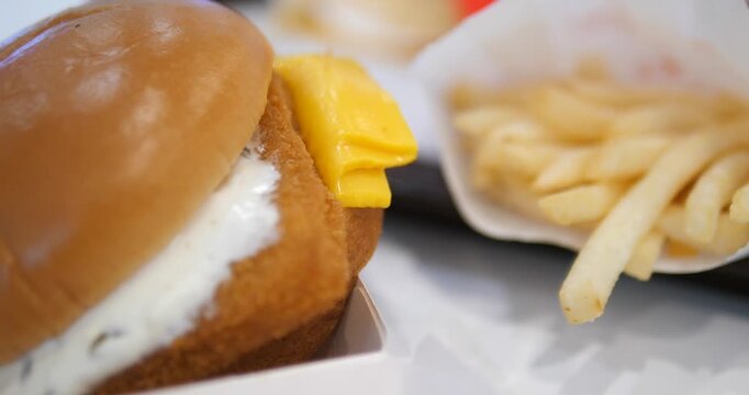 Close-up cinematic shot of a crispy fish burger with creamy tartar sauce and yellow cheese, appetizing fast food meal with fries background