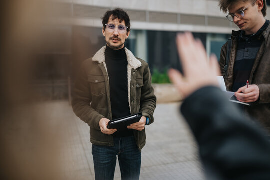 High school students stand outdoors while one holds a tablet and another takes notes. A candid, small group discussion captured in a casual urban setting.