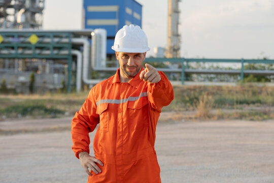 A confident industrial worker in an orange coverall and white helmet points forward at a processing plant or refinery.
