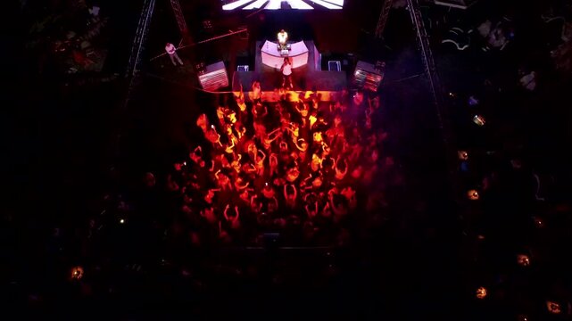 Drone aerial view captures outdoor night disco party crowd dancing around DJ stage. Energetic open air scene shows light beams spotlights pulsing wild music vibes useful lifestyle context.