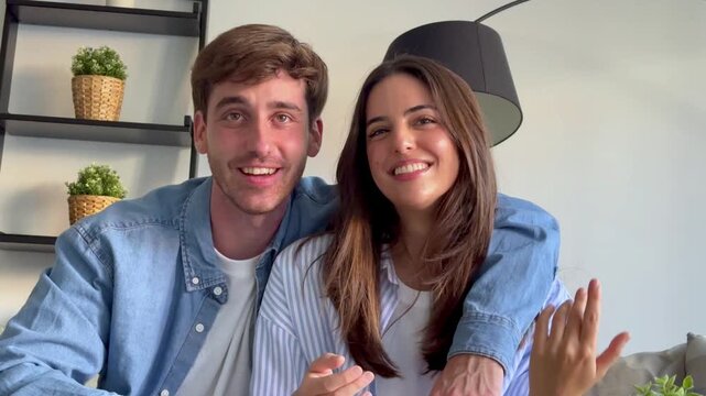 Cheerful young couple sitting at home, smiling and waving at the camera while engaging in a lively conversation during a video call with friends or family, sharing positive and happy emotions