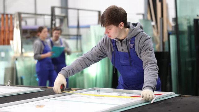 Young guy worker in uniform measuring glass sheet with tape measure on table in glass factory 